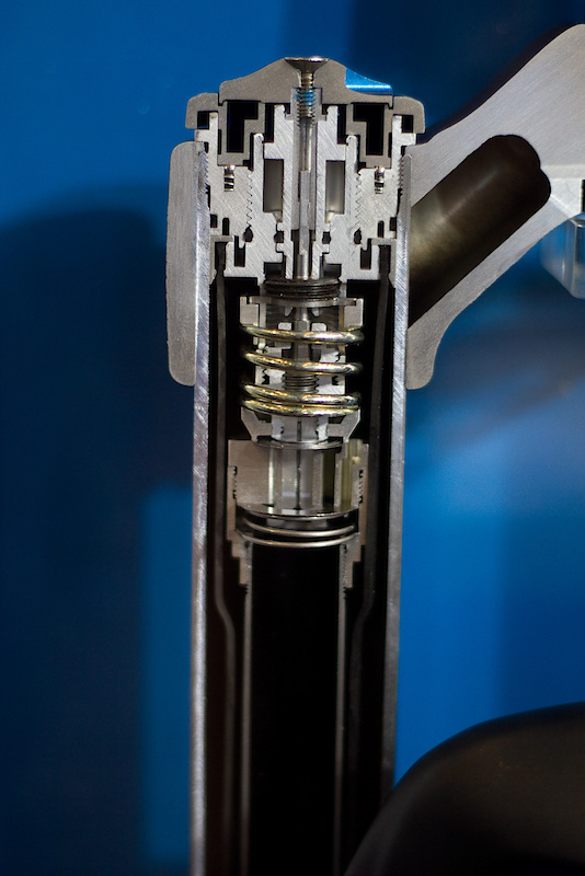 Fox had this very cool cutaway on display showing the insides of the FIT RC2 cartridge that is used in some of their 36 180 mm and 160 mm forks. For 2011 the new cartridge is inverted from the previous year, with the compression assembly and bladder now residing at the top of the leg and the rebound being found at the bottom. While this photo makes the internals look quite complicated - which of course they are - once you get a handle on the technology it will become much more clear. If you look closely at the photo above you should be able to spot the rubber bladder which sits just inside of the stanchion tube walls. The bladder serves to compensate for the oil displaced by the damping rod entering the cartridge. Because there is close to zero air within the oil filled cartridge, there needs to be space for the extra volume to go to as the rod enters the cart, which the rubber bladder accomplishes by expanding. The large gold colored spring at the center of the fork is the high speed compression spring. Turning the H/S dial atop the fork leg preloads this spring which holds down the shims that control the high speed oil flow. The more preload on the shims, the higher the oil force needed to flex them and the more high speed compression damping you'll have. Moving further down into the fork you may notice a second smaller diameter spring that is holding a single large diameter shim in place. This acts as the compression check valve, letting oil flow freely during the rebound stroke, but forcing the same oil through the compression assembly when the fork is being compressed. Not shown in this photo is the fork's hydraulic bottom out which is found on the coil sprung models, but absent on the air versions due to the air spring's inherent rising rate as the fork is compressed and volume decreases.